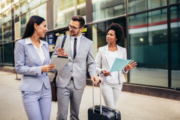 Three young business people talking to each other while walking outdoors with suitcase.