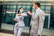 © Mediteraneo - Business man and woman with safety masks greeting with elbow bump in front of office building.