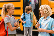 Two small classmates schoolchildren pupils talking discussing communicating with each other at the school yard getting off the school bus, going back to classes lessons after summer holidays lockdown.