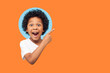 © khosrork - Wow look advertise here! Portrait of amazed little boy with curly hair pointing to empty place on background showing copy space for promotional ad. indoor isolated in a round hole on orange background