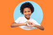 © khosrork - Portrait of smiling little boy with curly hair in white T-shirt showing heart shape with fingers, expressing innocent childhood love. indoor studio shot isolated in a round hole on orange background