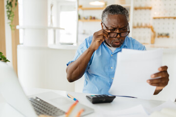 Amazed African pensioner sitting at home and looking at bills he has to pay. He is paying it online over a laptop.