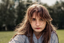 Casual Portrait Of Teenager Girl In A Park, Selective Focus. The Model Is With Light Make Up And Fake Freckles In Grey Clothes. Looking At The Viewer. Dark Green Trees In The Background