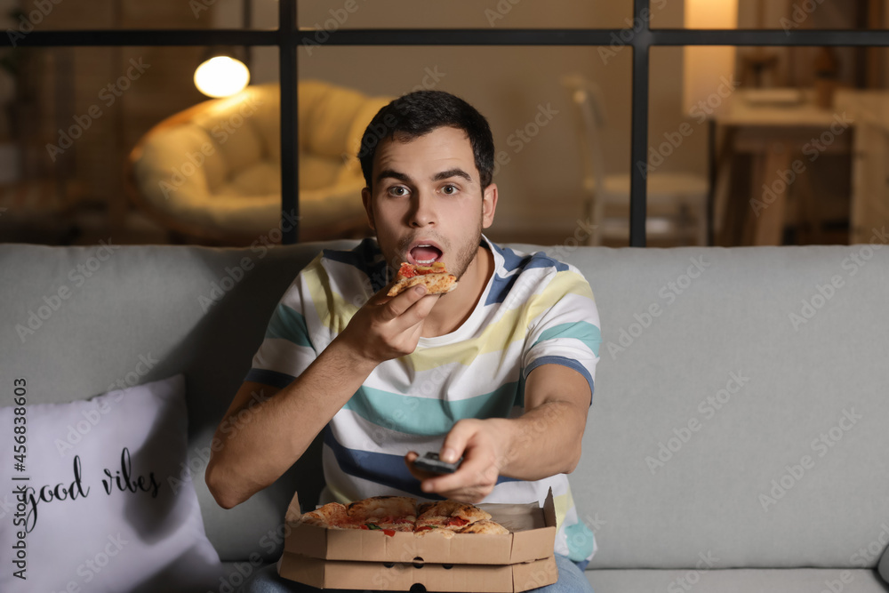 Handsome young man eating tasty pizza while watching TV at home in evening