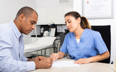 Young polite Latina cosmetologist doctor working with male client in clinic, filling out medical form before procedure ..