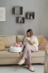 Wall Mural - Young woman with cup of tea and book sitting on couch by her baby daughter sleeping in small soft bed