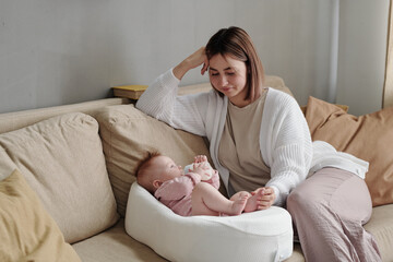 Wall Mural - Happy young mother sitting on couch next to her adorable baby daughter with bottle of milk