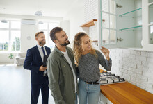 Boyfriend And Girlfriend Buying House. First Time Buyers Or Future Tenants Meeting Real Estate Agent And Looking Around New Home. Husband And Wife Looking At Good Quality Modern Wooden Kitchen Cabinet