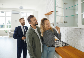 Boyfriend and girlfriend buying house. First time buyers or future tenants meeting real estate agent and looking around new home. Husband and wife looking at good quality modern wooden kitchen cabinet