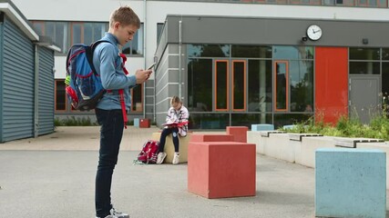 Wall Mural - Schoolboy standing with sell phone and schoolgirl sitting with book on background in schoolyard. 4K 60fps video