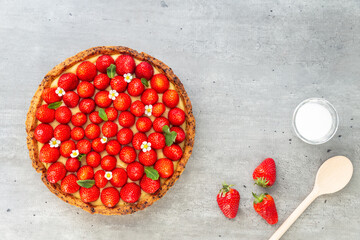 Wall Mural - French strawberry tart with wild strawberry leaves and flowers on top of a cooling rack with sugar and spoon on a grey background. Flat Lay, top view.