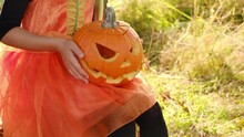 A Pumpkin With A Carved Evil Smile In The Hands Of A Child. The Halloween Holiday. Jacko' Lantern.Without A Face. A Girl In A Carnival Dress Is Sitting In The Dry Grass In Nature. Trick Or Treating 