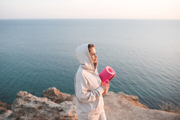 Wall Mural - Smiling beautiful woman wear hoody with hood  holding yoga mat posing over blue sea nature background outdoors. Adult girl training fitness at sunrise. Healthy active lifestyle. Sport concept. 20s.