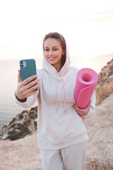 Wall Mural - Smiling adult girl 24-26 year old training wear beige hoodie sweatshirt holding yoga mat make photo with phone over sea nature background. Healthy lifestyle. 20s. Happy woman doing fitness on rock.