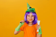 One Little Girl Childin A Carnival Costume Witch With Toothpaste And An Orange Brush For Halloween Is Isolated On A Yellow Background. Medicine, Dental Hygiene, Holidays Concept.