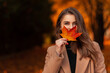 © alones - Happy beautiful woman in a fashionable beige coat and sweater covers her face with a colored autumn leaf in a park with orange fall foliage. Space for text