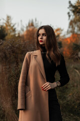 Wall Mural - Elegant beautiful young girl with red lips in a fashionable beige coat and sweater in the grass in nature. Female autumn style and beauty outdoors