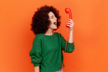 Side view of angry woman with Afro hairstyle wearing green casual style sweater screaming while talking to client in call center, negative emotions. Indoor studio shot isolated on orange background.