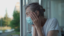 Worried Senior Woman In A Protective Medical Mask Sadly Looks Out The Window With Her Head Clasped In Her Hands In A Nursing Home. Depressed Lady At Home During The Covid-19 Pandemic