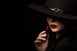 Beauty portrait of elegant woman with luxury lips makeup holding red lipstick. Model hides her face under wide hat, posing on black background. Close up studio portrait. Copy, empty space for text