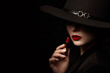 Beauty Portrait Of Elegant Woman With Luxury Lips Makeup Holding Red Lipstick. Model Hides Her Face Under Wide Hat, Posing On Black Background. Close Up Studio Portrait. Copy, Empty Space For Text