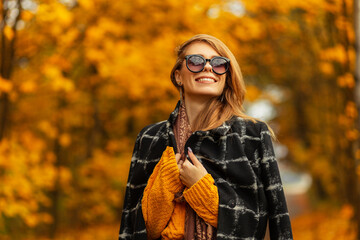 Happy beautiful young smiling woman with cute face in trendy black coat, knitted sweater, scarf and sunglasses enjoying a walk in autumn park with colored foliage. Fun emotions outdoors