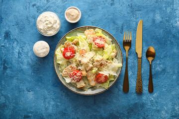 Plate of tasty Caesar salad and bowls with sauce on blue background