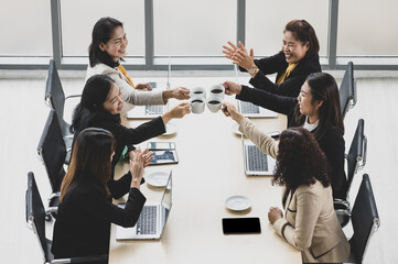 Wall Mural - High view of six businesswomen sitting together around wooden conference table , cheering and clinking coffee mug together with laptops and tablets on table in office. Concept for business meeting