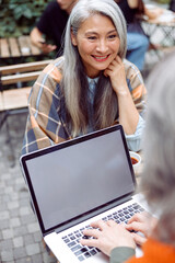 Wall Mural - Smiling senior Asian woman with friend using modern laptop sit together at small table on outdoors cafe terrace on nice autumn day