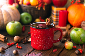 red mug with mulled wine. Cinnamon sticks stick out of the cup and a star of star anise floats. Fruits and spices are all around on a wooden table