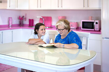 Elderly Woman Reading Book With Granddaughter In Light Kitchen At Home