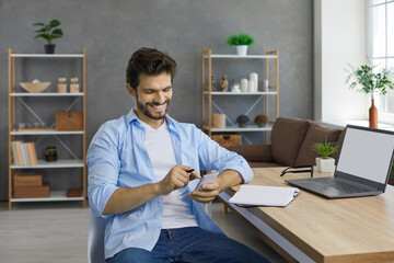 Happy handsome young man using mobile phone. Student in casual shirt sitting at working desk with laptop computer and paper notebook diary at home, holding cellphone, looking at screen and smiling