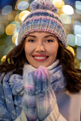 Wall Mural - winter holidays and people concept - portrait of happy smiling young woman in hat, mittens and scarf over christmas lights