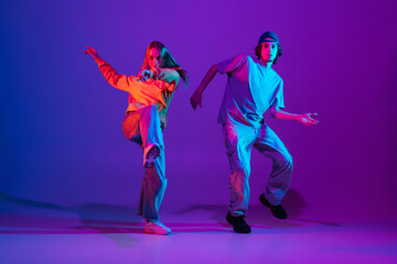 Wall Mural - Two dancers, young man and woman dancing hip-hop in casual sports youth clothes on gradient purple pink background at dance hall in neon light.