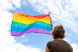 © aguscrespophoto - Unrecognizable woman on her back waving a rainbow flag. Looking at flag and sky on blue background with clouds.
