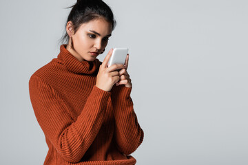 Wall Mural - young woman in knitted sweater messaging on smartphone isolated on grey