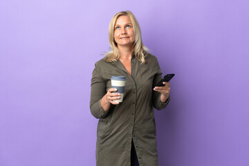 Wall Mural - Middle age Lithuanian woman isolated on purple background holding coffee to take away and a mobile while thinking something