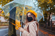 © maryviolet - Stylish young woman in mask against covid walks on autumn street under transparent umbrella during rain with leaves.