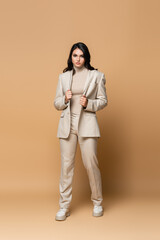 Sticker - full length of brunette young model in trendy suit adjusting blazer on beige