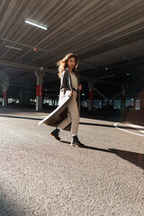 Wall Mural - Pretty young girl with curly hair in a fashionable long coat with boots walks on the street in the parking lot. Street feminine style and fashion