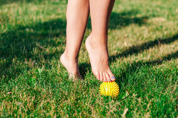 close-up of graceful bare feet of young woman standing on tiptoes on spiked massage ball on green lawn of courtyard summer, physiotherapy concept. 