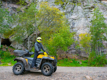 A Man On An ATV On A Rock Background. The Quad Bike Driver Looks Into The Distance. Travel By ATV. Nature Trip By Motorcycle Or Quad Bike. Concept Of Extreme Travel. Guy Tourist On An ATV.