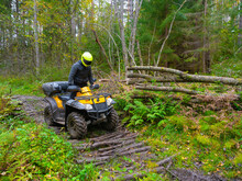 Man In A Yellow Helmet On An ATV. He Rides His ATV Through The Forest. Off-road Driving Concept. ATV Driver Overcomes An Obstacle On The Road. Quad Bike As A Symbol Of Motorcycle Racing