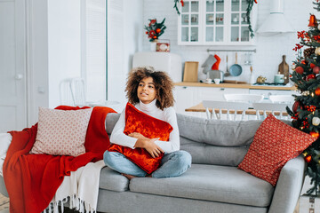 Wall Mural - Kinky haired lady with dark skin wearing white sweater holding red pillow on head sits in lotus yoga pose on sofa closeup