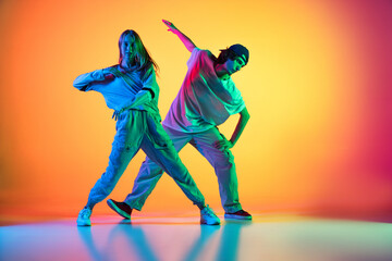 Two dancers, young girl and boy dancing hip-hop in casual sports youth clothes on gradient multi colored background at dance hall in neon light.