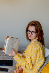 Young woman in comfortable yellow pajamas works while lying the bed in the bedroom. Business woman maintains a social distancing works remotely. New normal concept