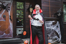Happy Mature Man In Vampire Halloween Costume Standing On Decorated Porch With Pape Cut Bat