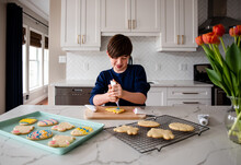 Young Boy Decorating Easter Cookies On Counter Of A Modern Kitchen.