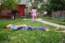 A Boy Lays In Green Grass In Yard With Sister And Cat In Background