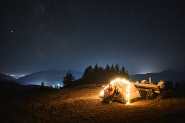 Wall Mural - Couple travelers sitting inside illuminated camp tent and looking at evening sky with stars. Magnificent view of blue starry sky over mountain valley. Concept of travelling, hiking, night camping.